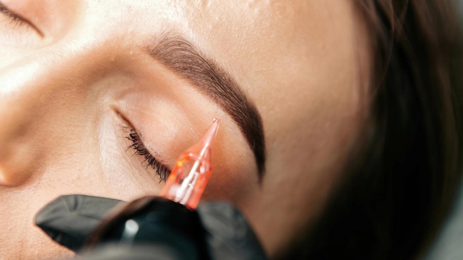 Nahaufnahme einer Permanent-Make-up-Künstlerin, die mit einem Präzisionswerkzeug eine Nachbesserung der Augenbrauen an einem geschlossenen Augenlid durchführt